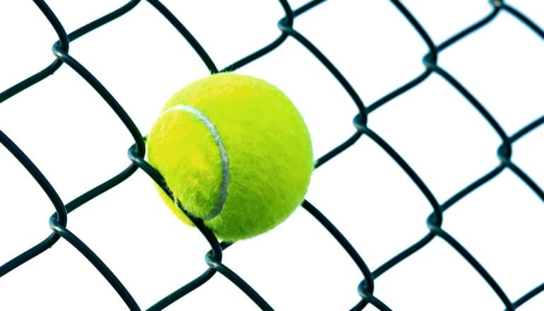 A tennis ball is stuck in a chain-link fence with white in the background