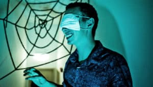 A man with a blindfold on reaches out in a darkly lighted haunted house with a spider web in the background