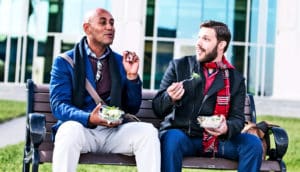 Two people talking on a bench while eating lunch outside an office building