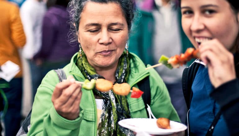 Two women eat skewers of vegetables at a food festival