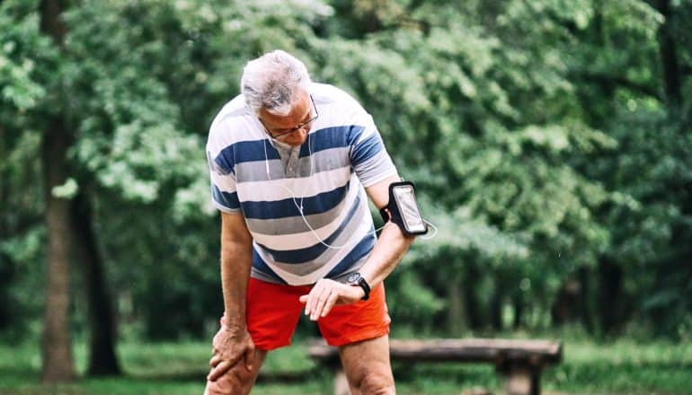 An older man in orange shorts and a striped t-shirt looks down at his activity tracker, with green trees in the background