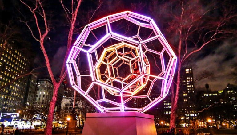 A pink, light-up structure that looks just like a buckyball sits on a pedestal in a big city