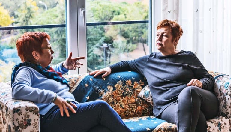 Two women with red hair, one in blue and one in black, sit on a couch and discuss something intently