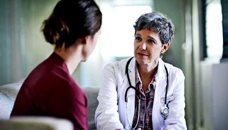 A doctor in a white coat with short hair talks to a patient as they both sit on the couch