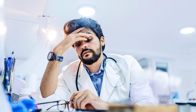 A doctor sitting at a desk in a white coat over a blue shirt rubs his eyes in frustration while holding his glasses in his other hand