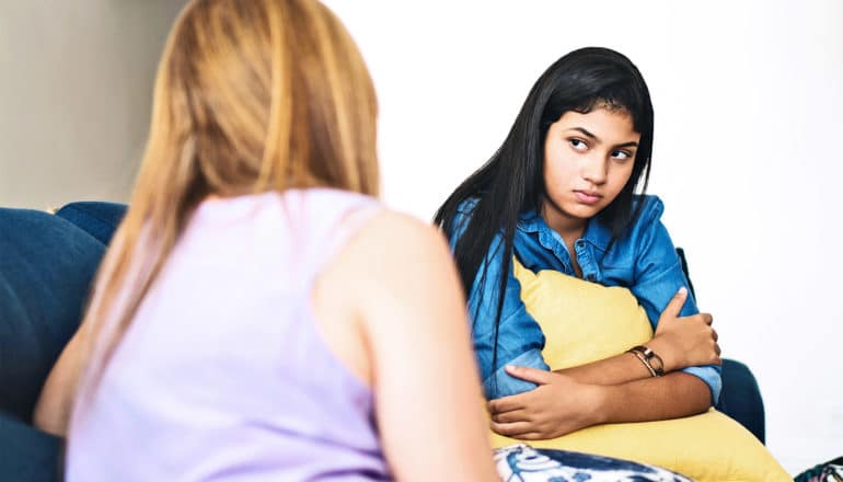 A mother in a purple top speaks with her daughter, whose wearing denim and is looking at her mother while holding a yellow pillow