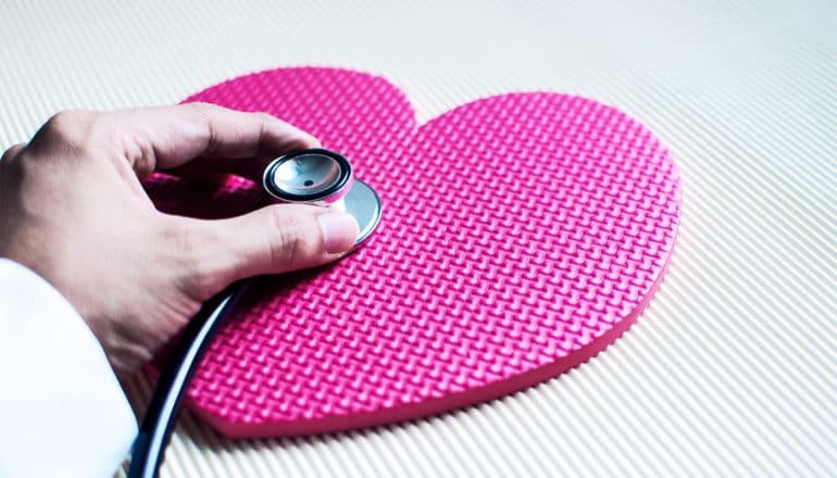 A doctor uses a stethoscope on a flat, pink/red cutout of a heart