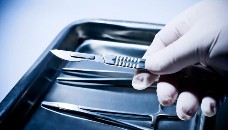 A gloved hand picks up a scalpel from a metal dish of surgical implements