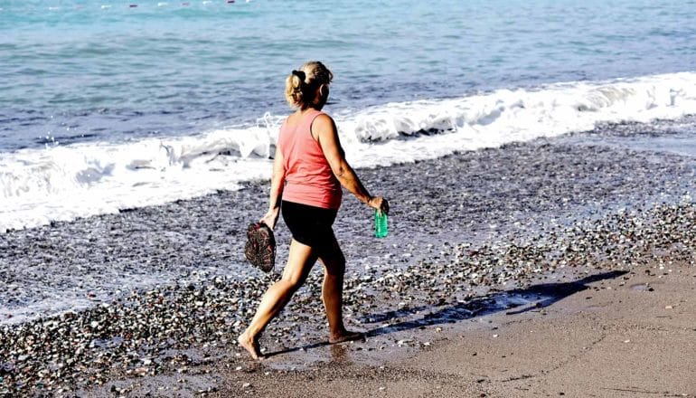 An older woman in a pink top holds her shoes and a waterbottle as she walks on a rocky beach, with a small wave crashing behind her