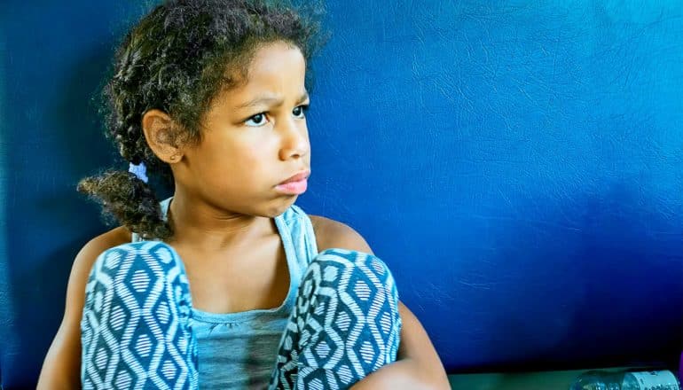 A young girl in blue sits on a blue bench seat with a puzzled or disappointed look on her face