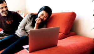 A woman on an orange couch doubles over laughing at a laptop screen as a man laughs beside her in the background