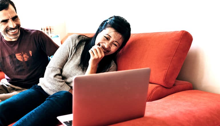 A woman on an orange couch doubles over laughing at a laptop screen as a man laughs beside her in the background