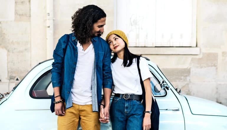 A couple stands together holding hands outside a small car parked against a wall, as they both look at each other. The man is wearing yellow pants, a white shirt, and a blue overshirt. The woman is wearing a yellow beanie, white top, and blue pants.