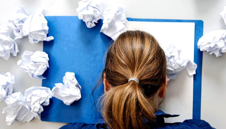 A woman has her head on her desk, which is covered in crumpled paper