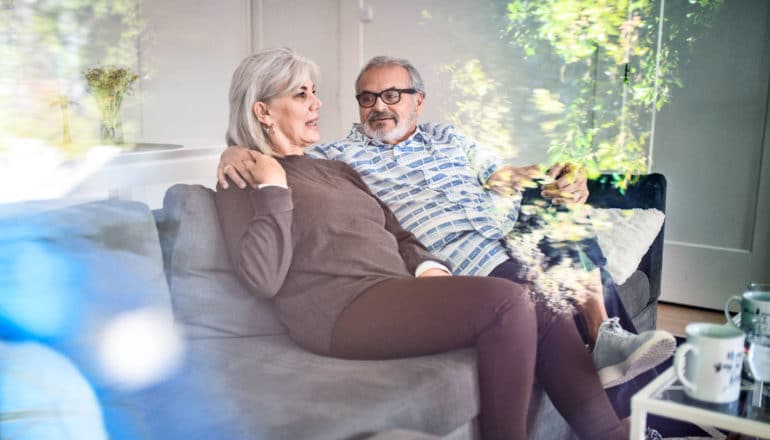 middle aged couple talk on couch, his arm around her shoulders