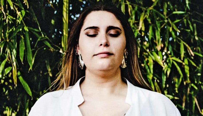 A woman in white is meditating with some leaves in the background behind her