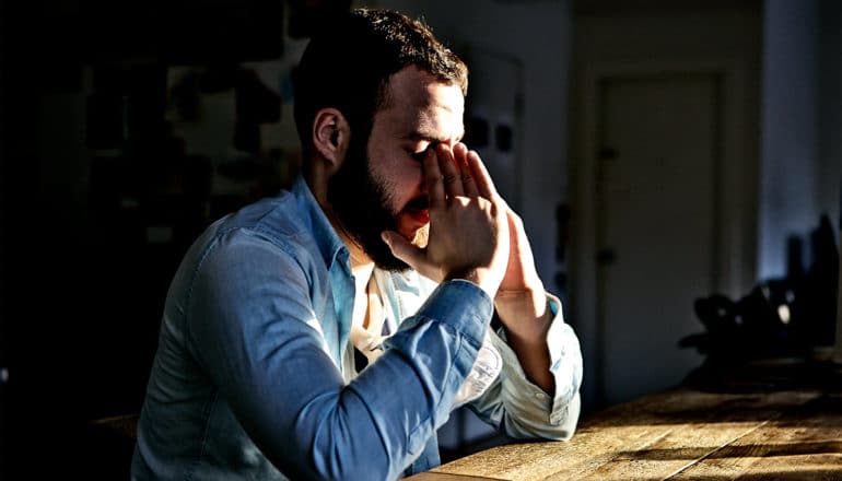 A man in a blue shirt sits in a dark room at a table with his hands touching his eyes and light coming through a window