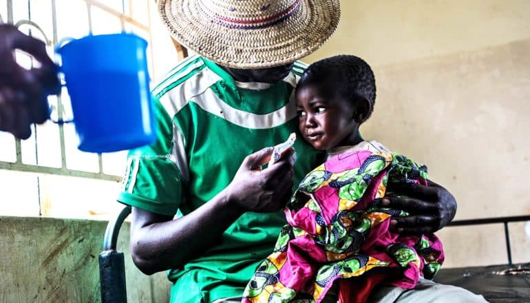 A father in a green shirt and wide-brimmed hat feeds his crying daughter, bundled in a brightly colored blanket