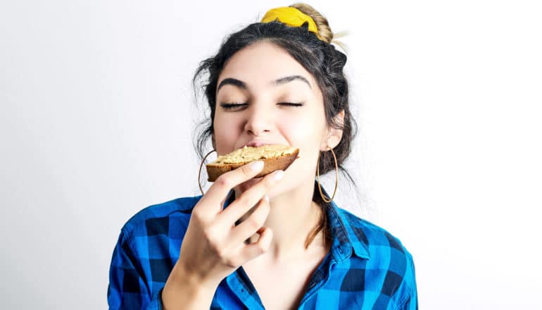 A woman in a blue button-up shirt takes a big bite of some bread with peanut butter on it, closing her eyes in enjoyment