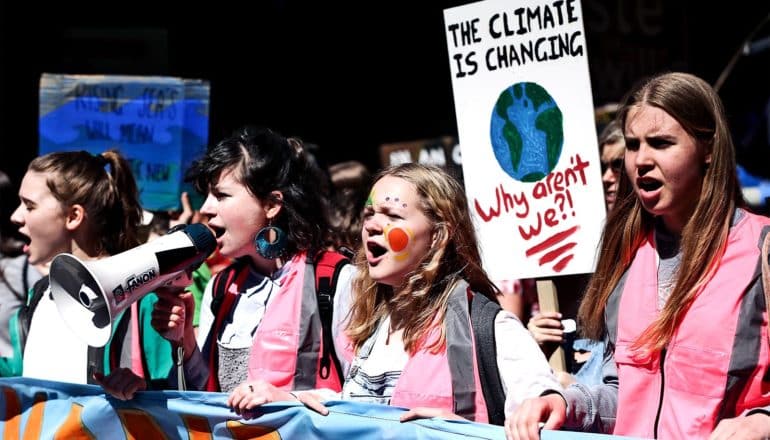 4 young women protest for climate action, all in pink vests. One holds a megaphone, another a sign that reads "The climate is changing. Why aren't we?"