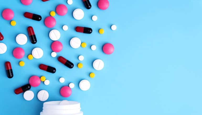 Many different colored pills (some pink, some black and red, some yellow, some white) burst out of a white pill bottle, spilling onto the blue background