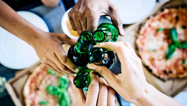 four people clink green beer bottles over some pizzas