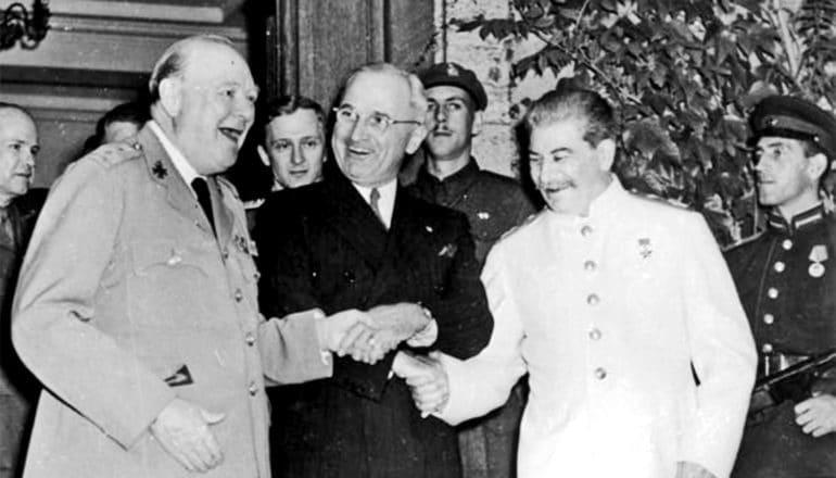 Churchill looks to be saying something as he, Truman, and Stalin, who is dressed entirely in white, shake hands together in a triple shake, with Churchill and Stalin on either side of Truman