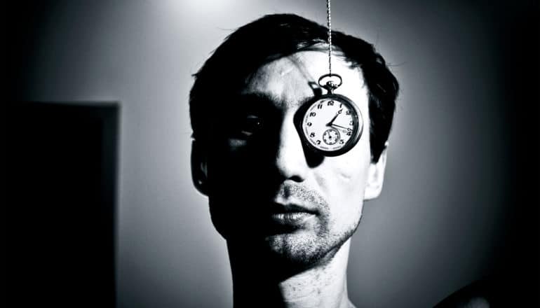 A man's face is half-covered in shadow as a pocket watch hangs down over his left eye