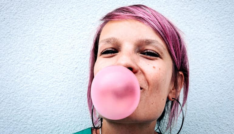 A young woman with pink hair blows a pink bubble with chewing gum while standing against a white wall