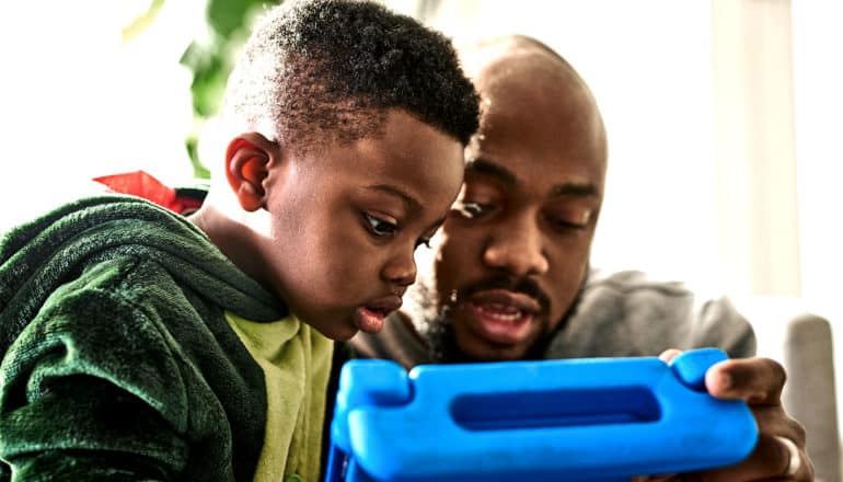 A father reads to his son from a tablet in a bright blue case. The son is wearing pajamas that double as a dinosaur costume