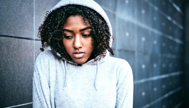 A young woman walks near a dark wall in a light hoodie pulled up over her head