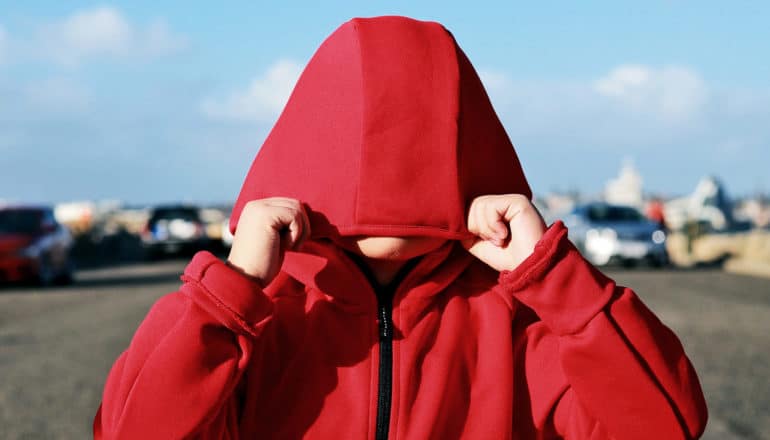 A young child pulls a red hoodie down over his face with a road and blue sky in the background