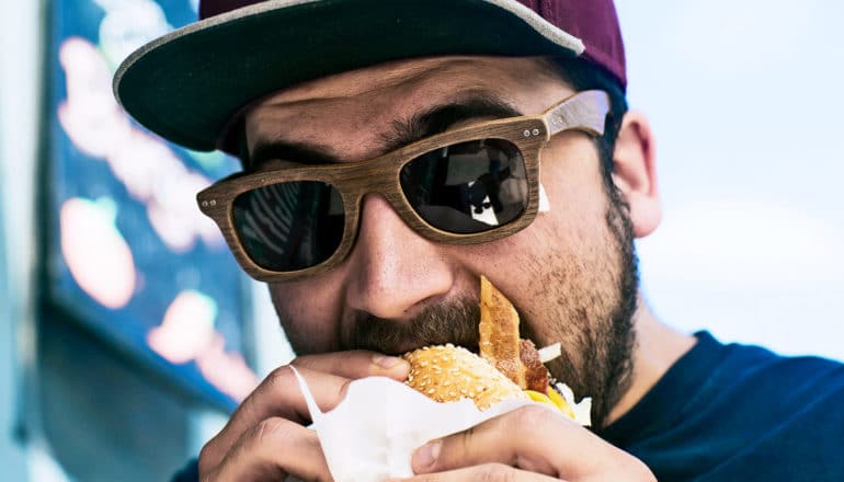 A man with a beard, hat, and sunglasses takes a bite of a burger