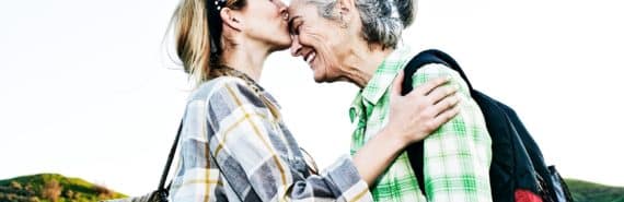 A young woman wearing a flannel shirt and a backpack kisses an older woman on the forehead as she smiles, with mountains and sky behind them