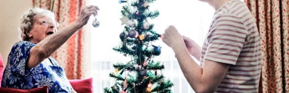 elderly person holds up ornament while decorating small christmas tree with younger person