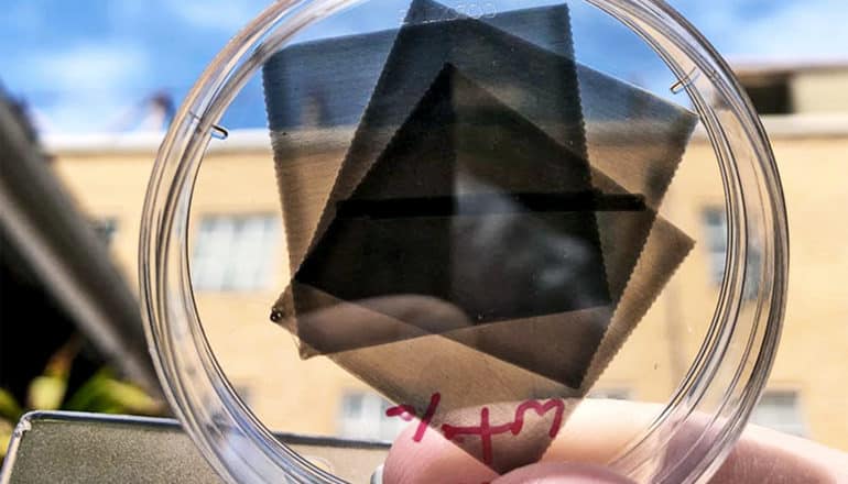 A few dark, transparent squares of the polymer sit on a petri dish a researcher is holding up to the sky, with a building visible behind the polymer sheets