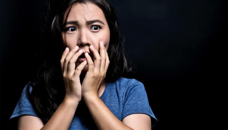 A woman in a blue t-shirt looks horrified against a black background