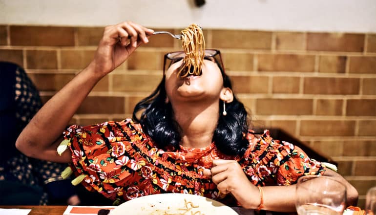 A young woman in an orange dress leans her head back to eat a huge forkful of noodles with a brick wall in the background