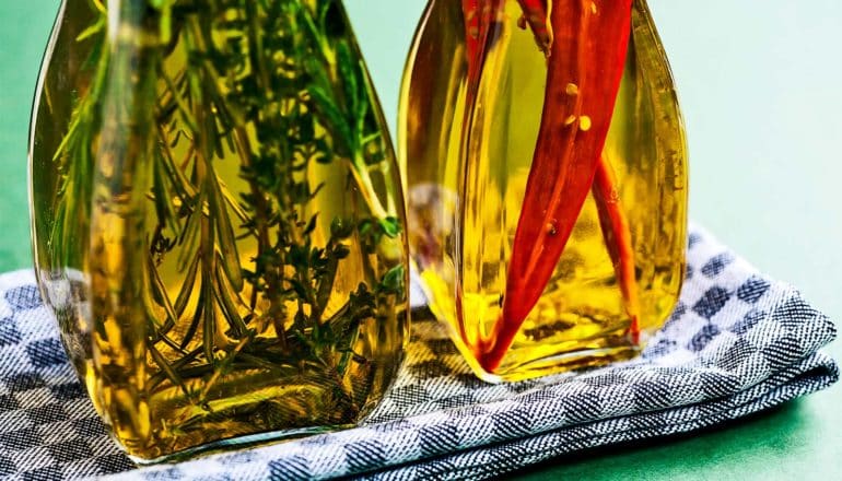 two bottles of infused olive oil sit on a checkered tea towel, one bottle with herbs and the other with spicy red peppers