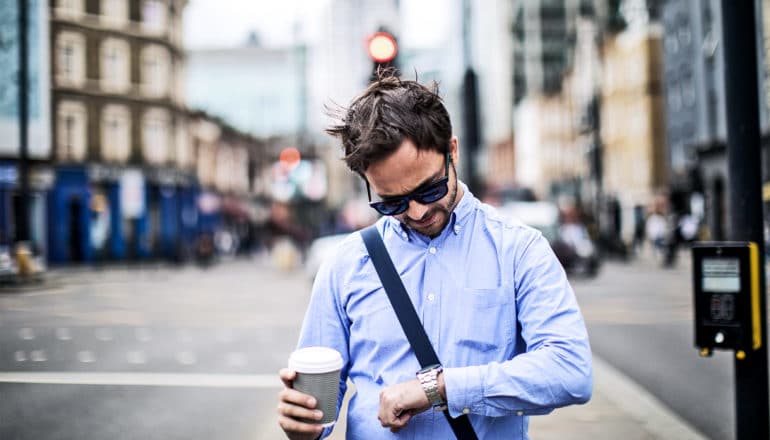 A man in a blue shirt and sunglasses holds a coffee cup as he looks at his watch while walking down a city street.