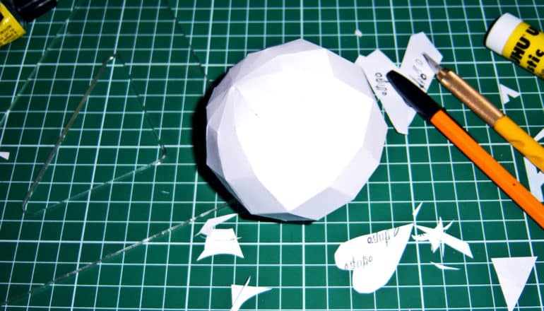 A kirigami sphere sits on a green work surface with pens, rulers, and scraps of paper around it