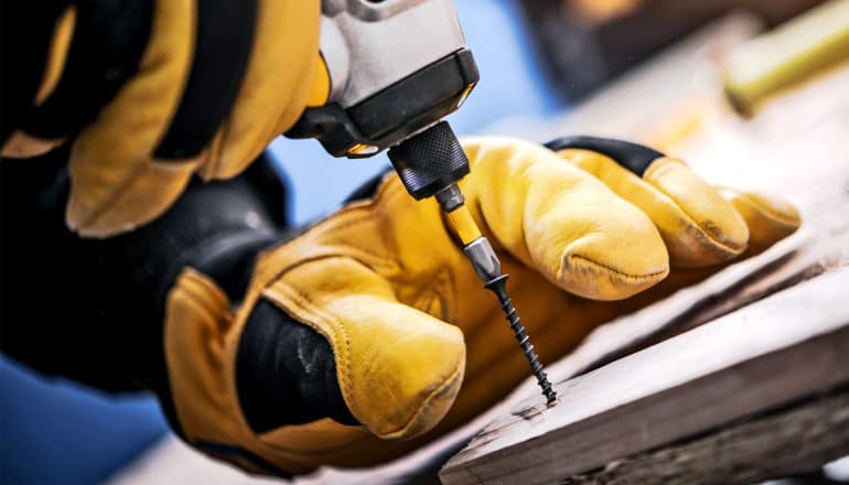 A person in yellow and black gloves uses a drill to screw in a nail