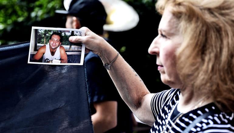 An older woman in a black and white striped t-shirt holds up a photograph of her son as a man with a tuba walks past her