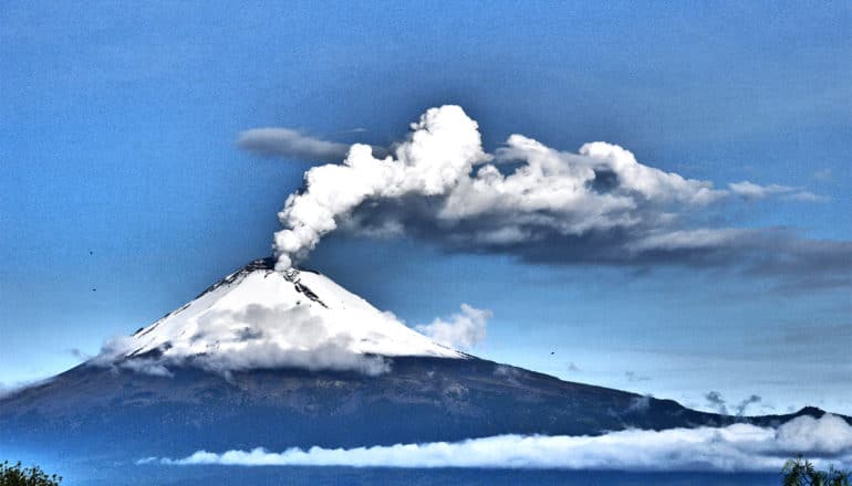 A snow-covered volcano spews smoke against a bright blue sky