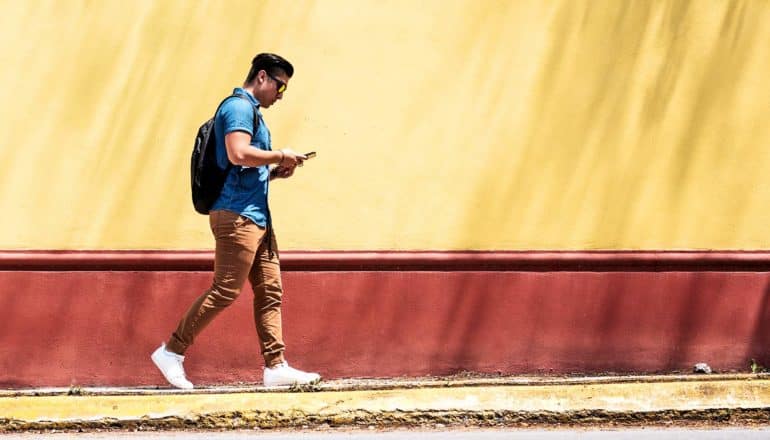 A man in a blue shirt, khaki pants, and white shoes walks near a red and yellow wall while looking down at his phone