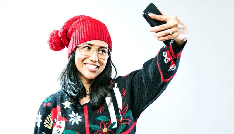 A woman in a red beanie with a pom-pom on top and a Christmas sweater holds up her phone to take a selfie