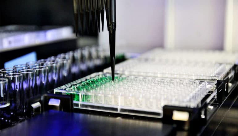 A robotic pipette places green liquid in one of many plastic wells