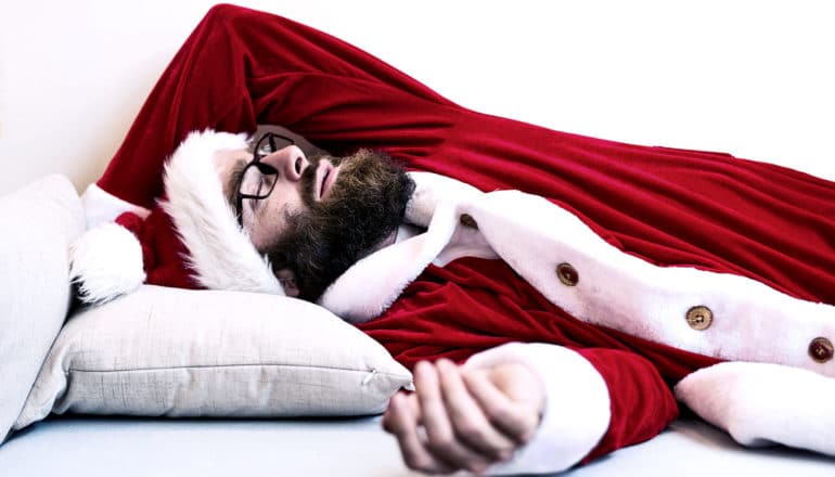 A bearded man in a red Santa costume with white fur lining sleeps on a couch