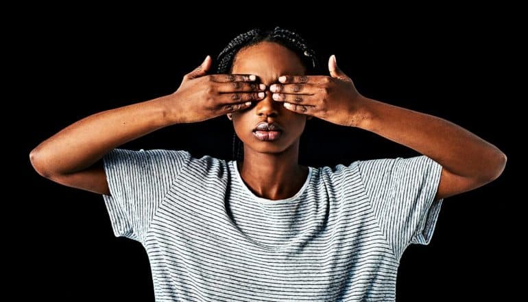 A woman in a white and black striped t-shirt covers her eyes with her hands against a black background