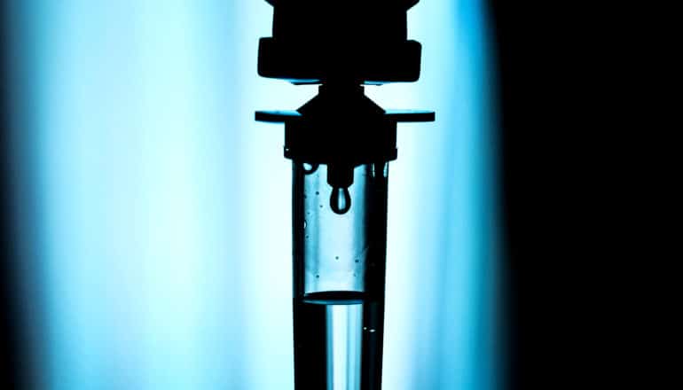 A IV drip in silhouette against light from a window, with a single drop about to fall